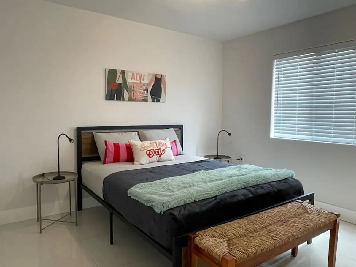 The image features a stylish, sunlit bedroom with a plush queen bed adorned with decorative pillows, modern wall art, chic nightstands, and a cozy bench, creating a welcoming and comfortable atmosphere.