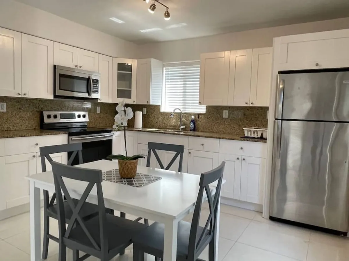 A bright, welcoming kitchen with a central island, stainless steel appliances, white cabinetry, and sunlight streaming through a nearby window. The layout is open and inviting, with generous counter space perfect for preparing meals or entertaining guests. The kitchen seamlessly connects to an adjacent dining area, visible in the background, supporting communal cooking and social gatherings.