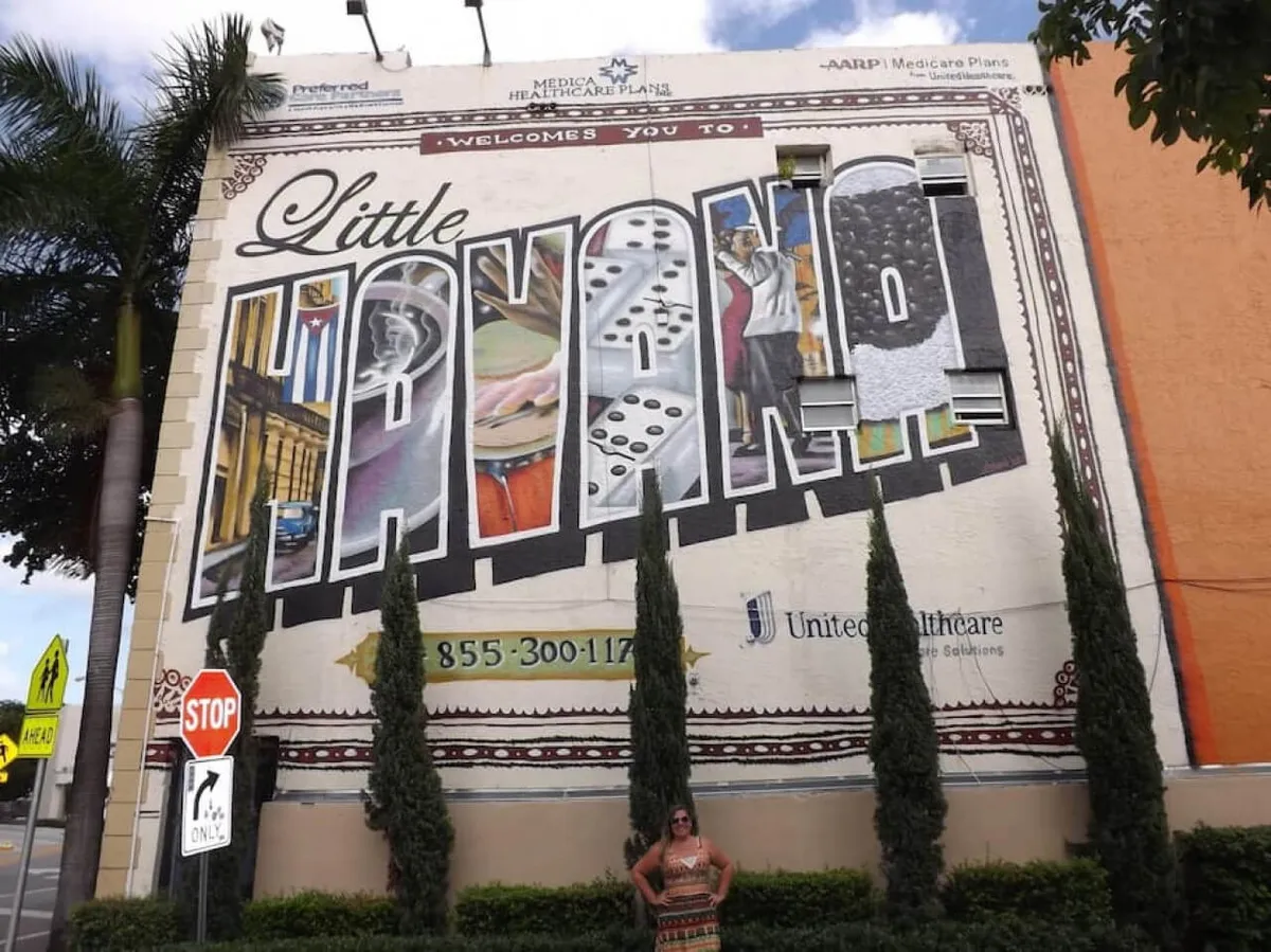A vibrant street scene featuring a large mural that reads 'Little Havana,' flanked by tall palm trees and a stop sign. A person stands in front of the mural, giving life and scale to the artwork. The atmosphere is lively, with a celebratory mood that captures the energy of the neighborhood and its cultural pride.