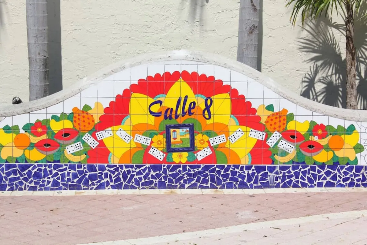 A close-up view of a mosaic mural adorned with vibrant fruit motifs and the inscription 'Calle 8.' The artwork bursts with color and reflects the joyful, festive spirit of local Cuban culture. The scene suggests a public space often visited for cultural events and art appreciation.