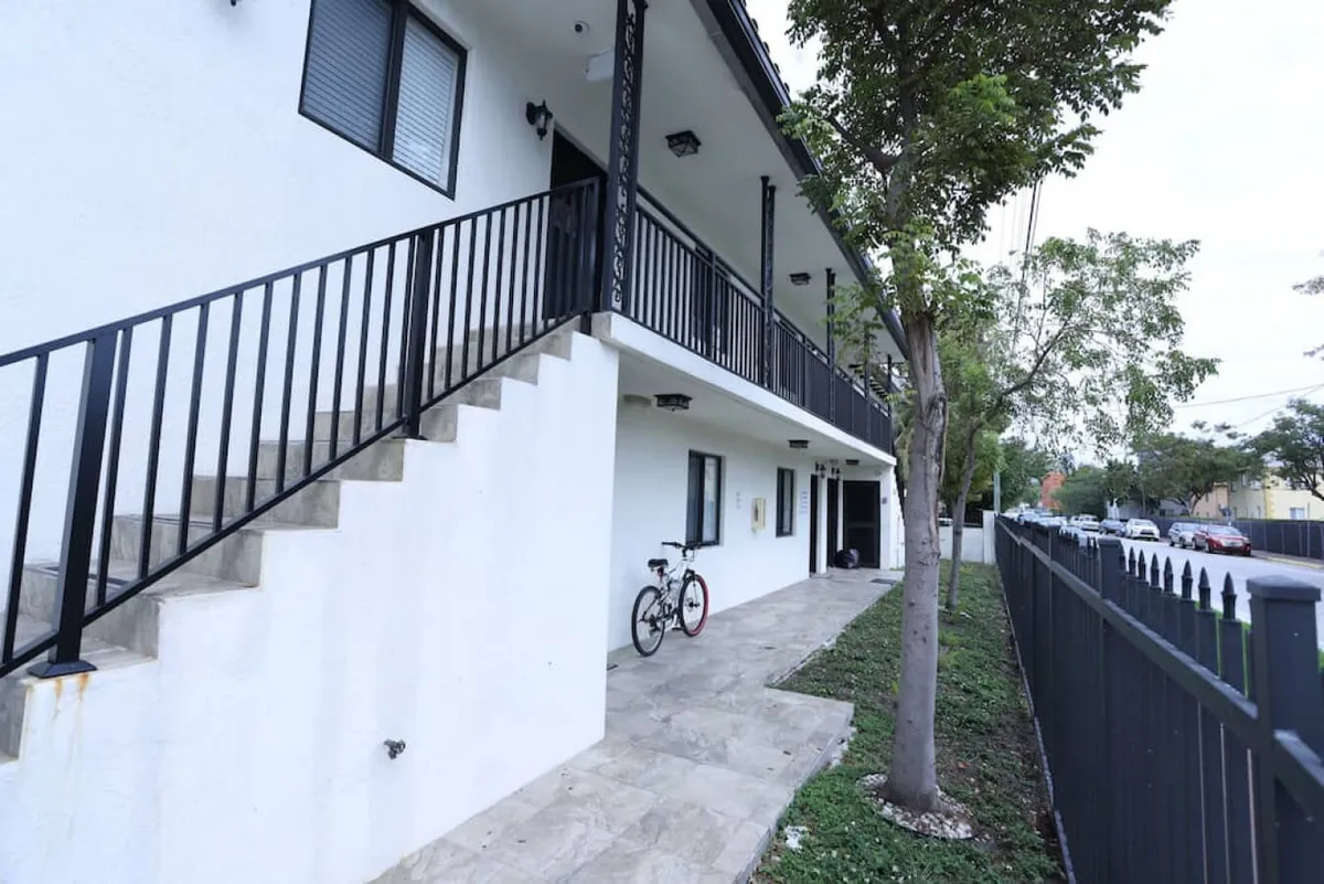 The image features the building's exterior focused on a staircase and landscaping, with a bicycle nearby and the entrance visible. The overall scene is welcoming and accessible, showcasing the approach to the unit.