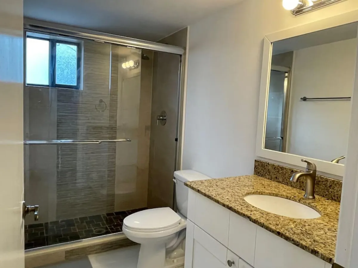 A fresh and modern bathroom with a glass shower enclosure, granite countertop, and large mirror. The space is bright and well-lit, ideal for morning routines or unwinding after a day out. Sleek fixtures and clean lines create a contemporary, inviting feel.