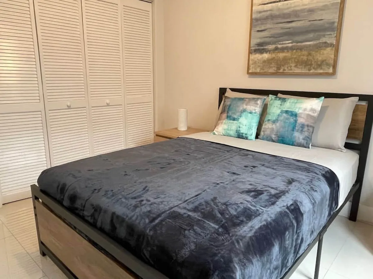 A second cozy bedroom with a queen bed draped in a dark blue plush blanket and accented with colorful pillows. Abstract artwork and gentle natural light add to the serene, inviting vibe. The room feels both modern and intimate, ideal for couples, friends, or families.