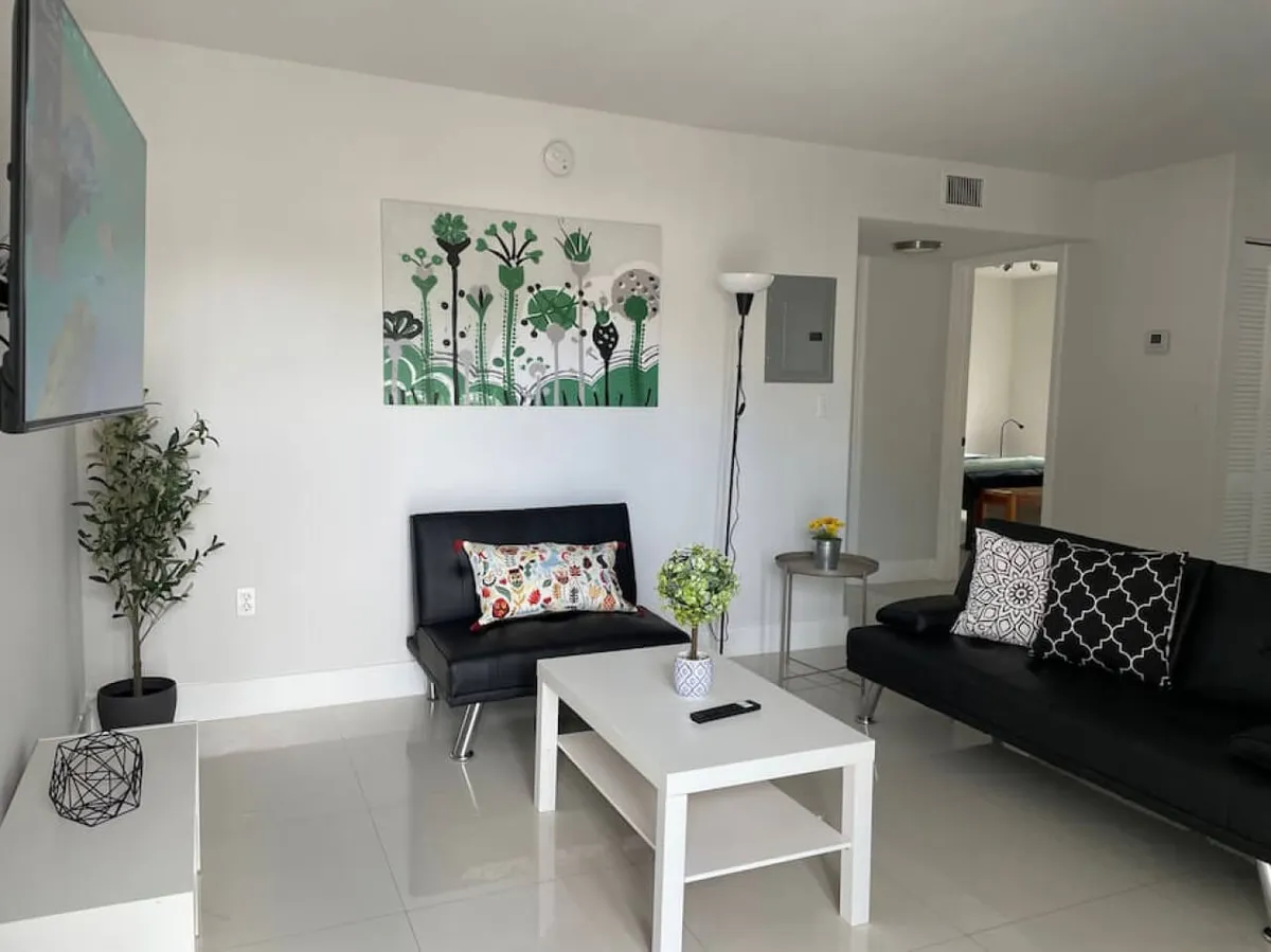 A bright, contemporary living room featuring black sofas adorned with green wall art, a central coffee table with decorative items, and ample space for relaxing or socializing. The arrangement allows for easy conversation and group gatherings, with natural light filtering through large windows.
