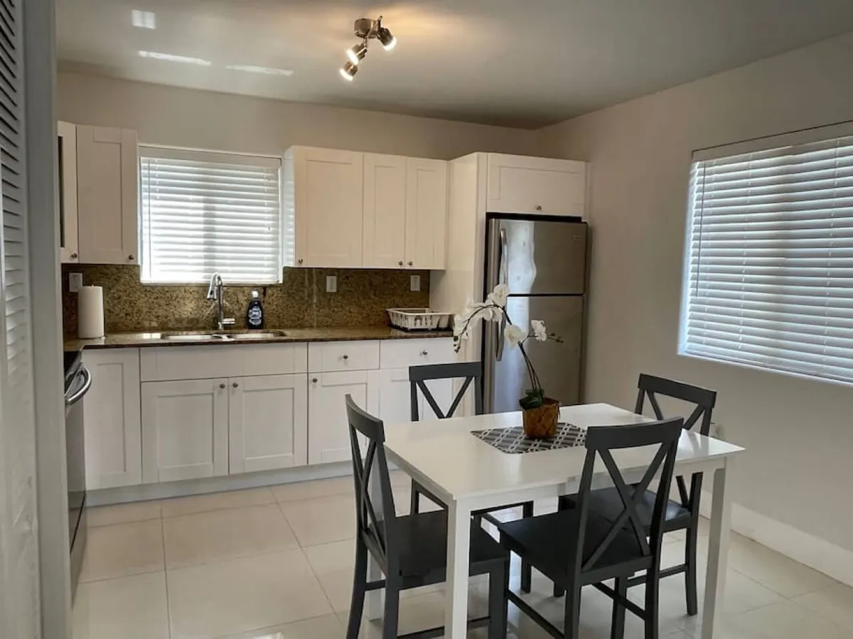 A view of the open kitchen and adjacent dining area, showcasing a kitchen island, a dining table, sleek stainless steel refrigerator, and granite countertops. The scene is set for cooking, socializing, and shared meals, with a warm and contemporary ambiance.